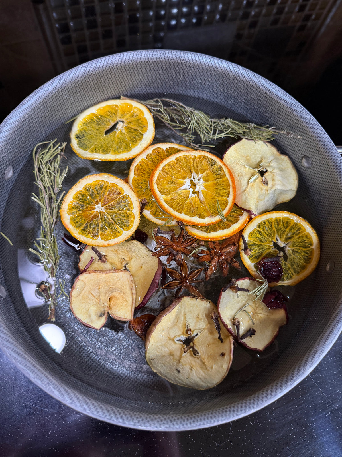 A close-up overhead shot of a metal pot containing dried citrus slices, apple slices, star anise, and rosemary floating in water, likely a home fragrance or tea infusion setup.