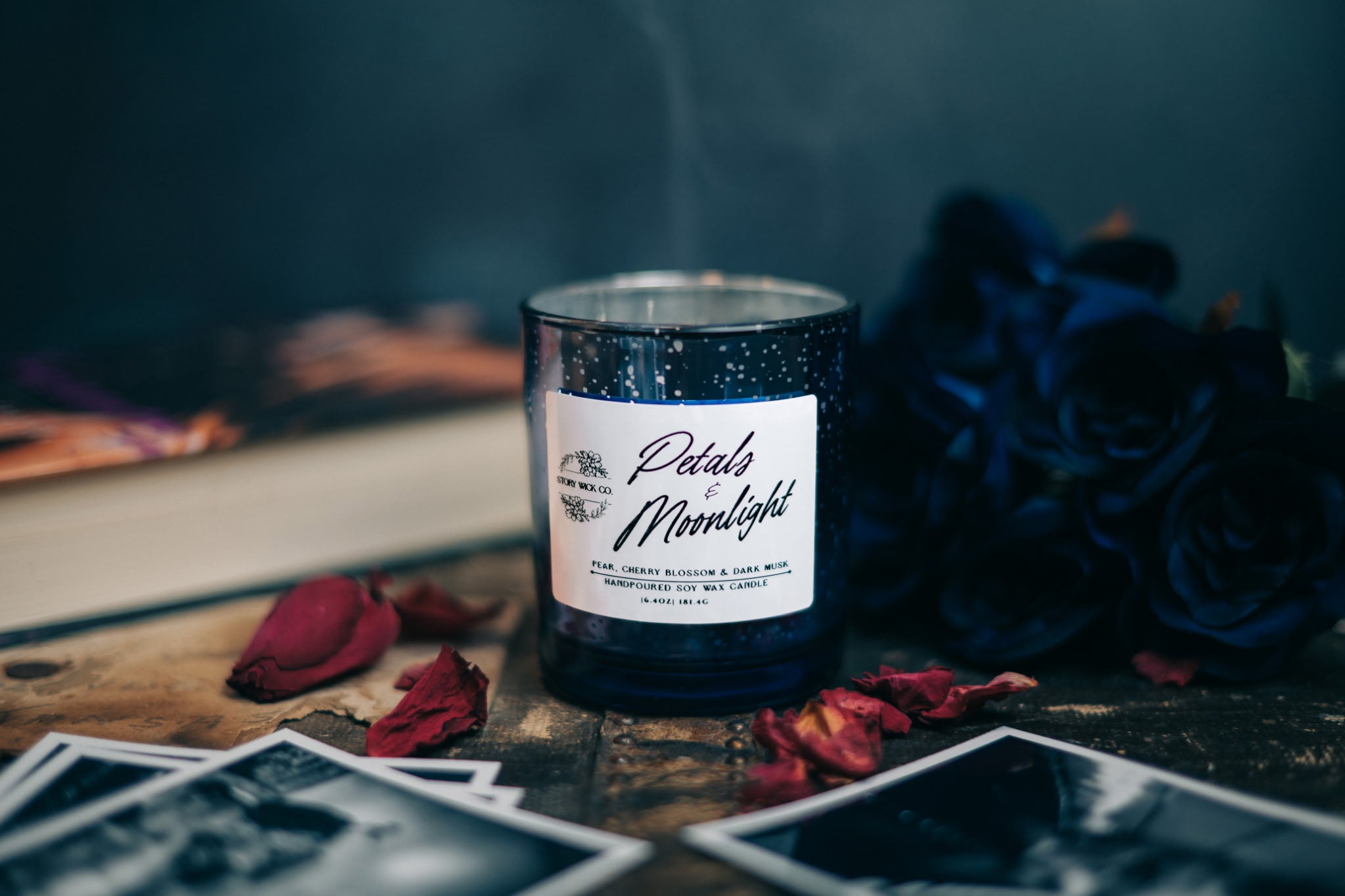 Candle labeled 'Petals of Moonlight' with flowers and black and white pictures on a dark surface