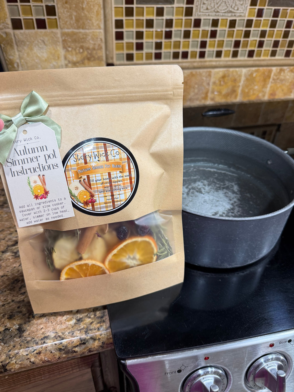 A kraft paper stand-up pouch labeled 'Autumn Summer Pot Instructions' with a clear window showing dried orange slices and herbs, placed on a kitchen counter beside a pot on a stovetop.