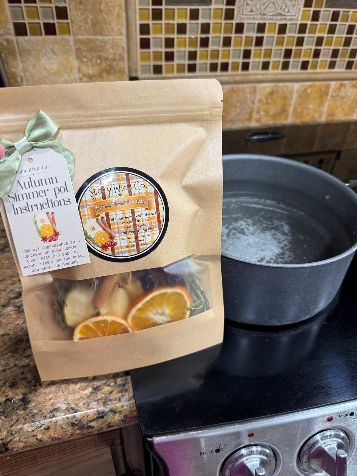 A kraft paper stand-up pouch labeled 'Autumn Summer Pot Instructions' with a clear window showing dried orange slices and herbs, placed on a kitchen counter beside a pot on a stovetop.