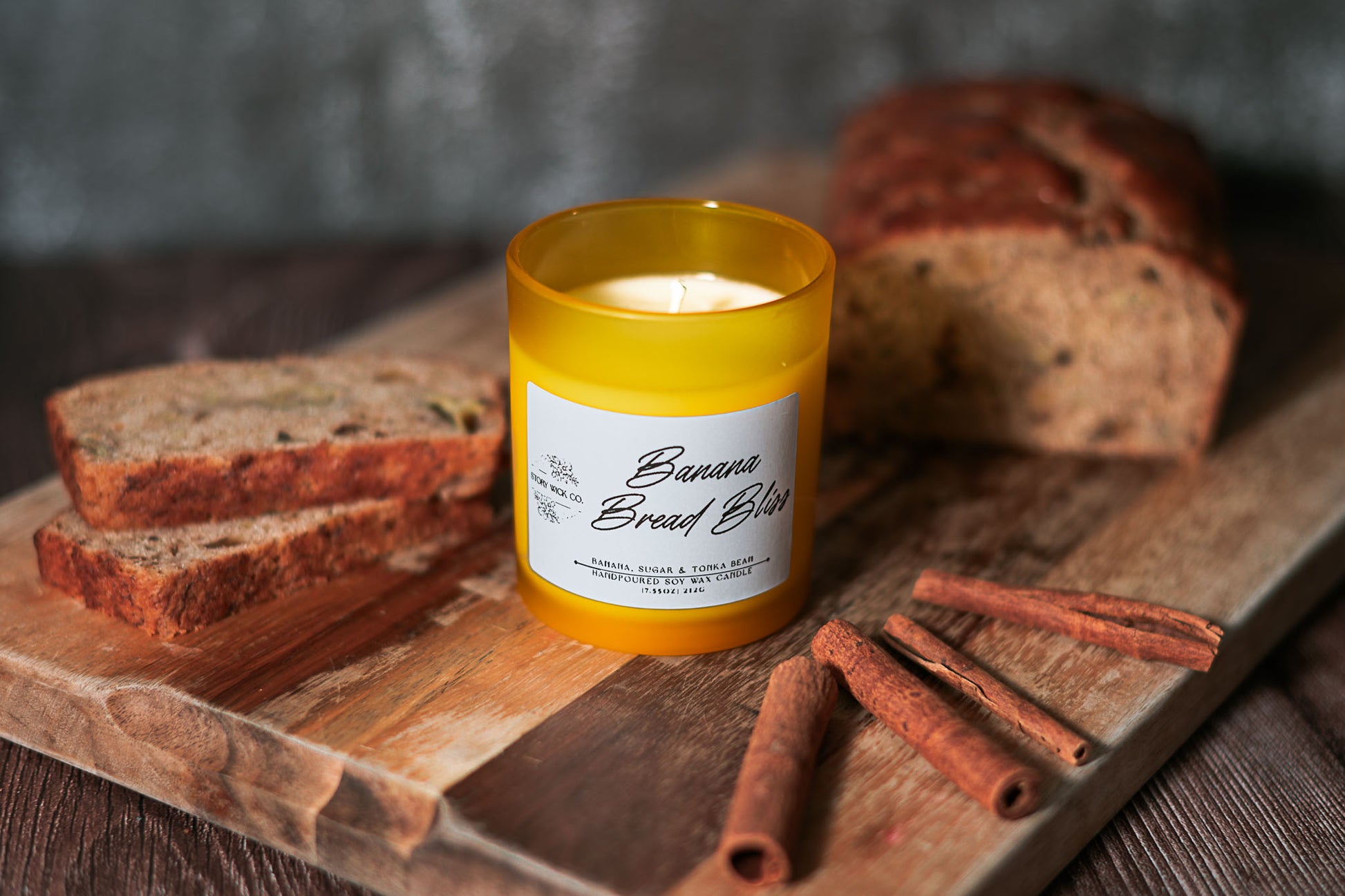 Candle labeled 'Banana Bread Bliss' on a wooden board with bread slices and cinnamon sticks.