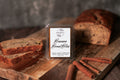 Packaged wax melt labeled 'Banana Bread Bliss' on a wooden surface with bread and cinnamon sticks.
