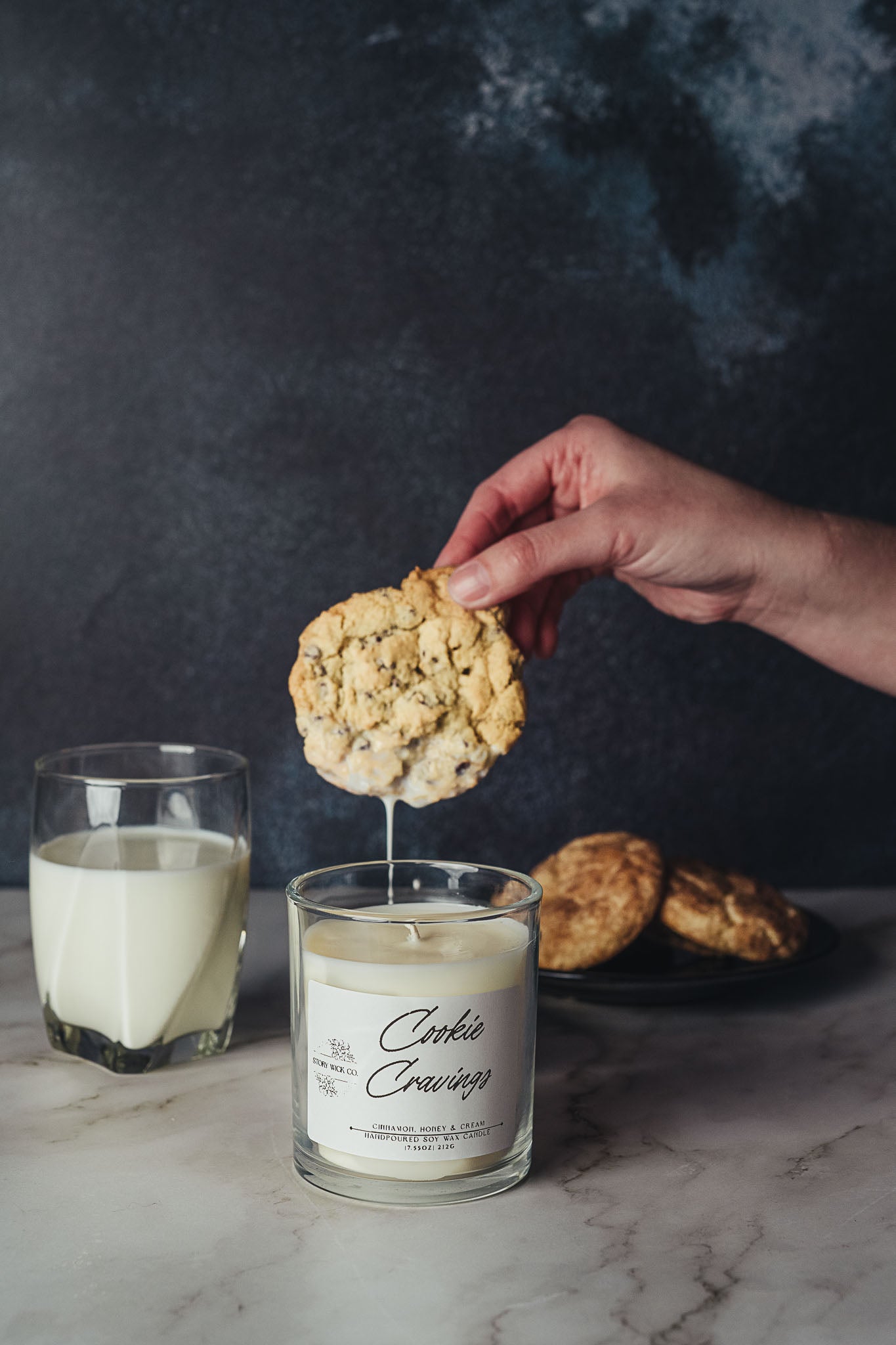 Person dipping a cookie into a candle labeled 'Cookie Cravings' with a glass of milk on a dark background.
