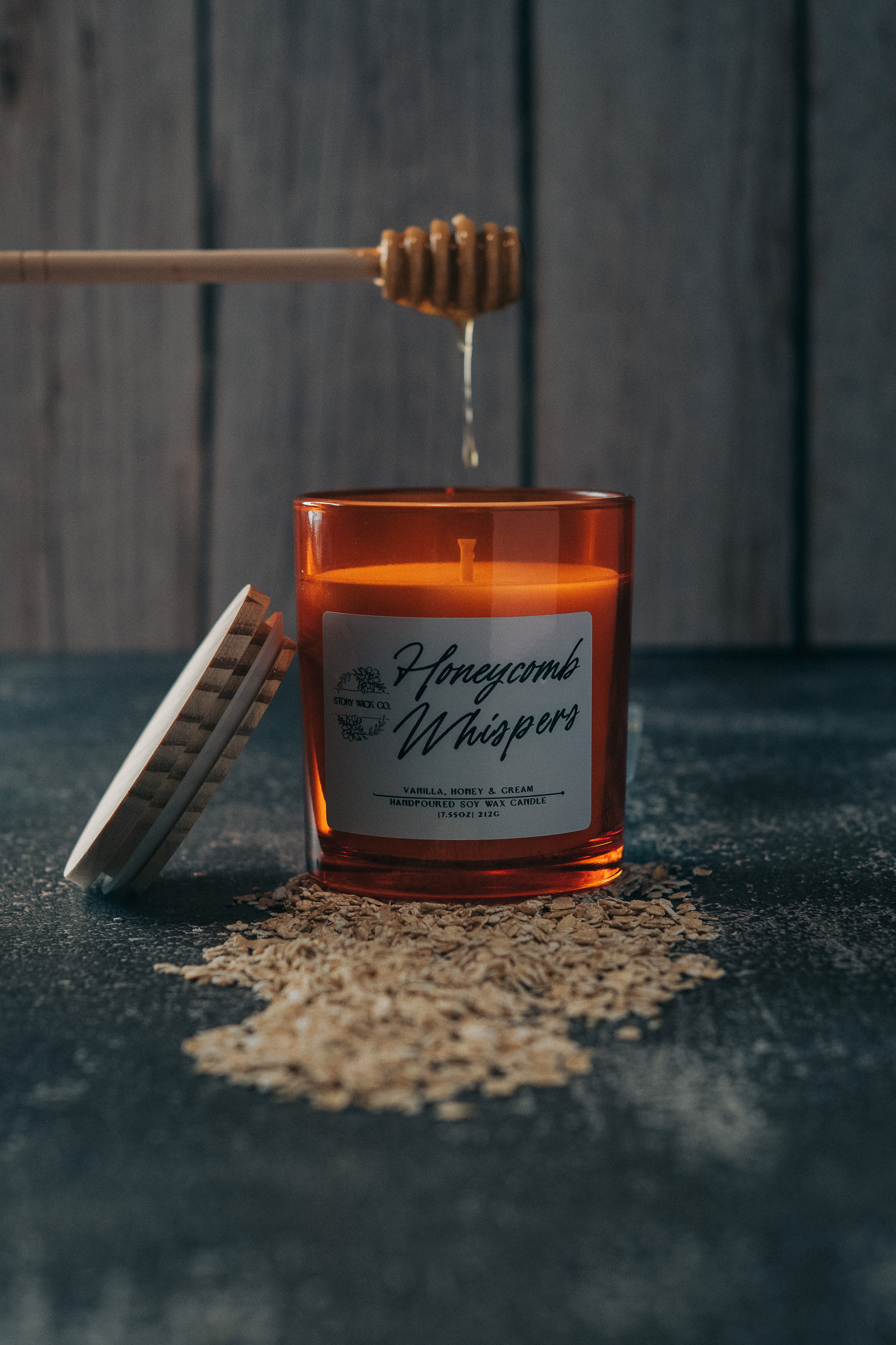 Candle labeled 'Honeycomb Whispers' with honey and a honey dipper on a dark surface.