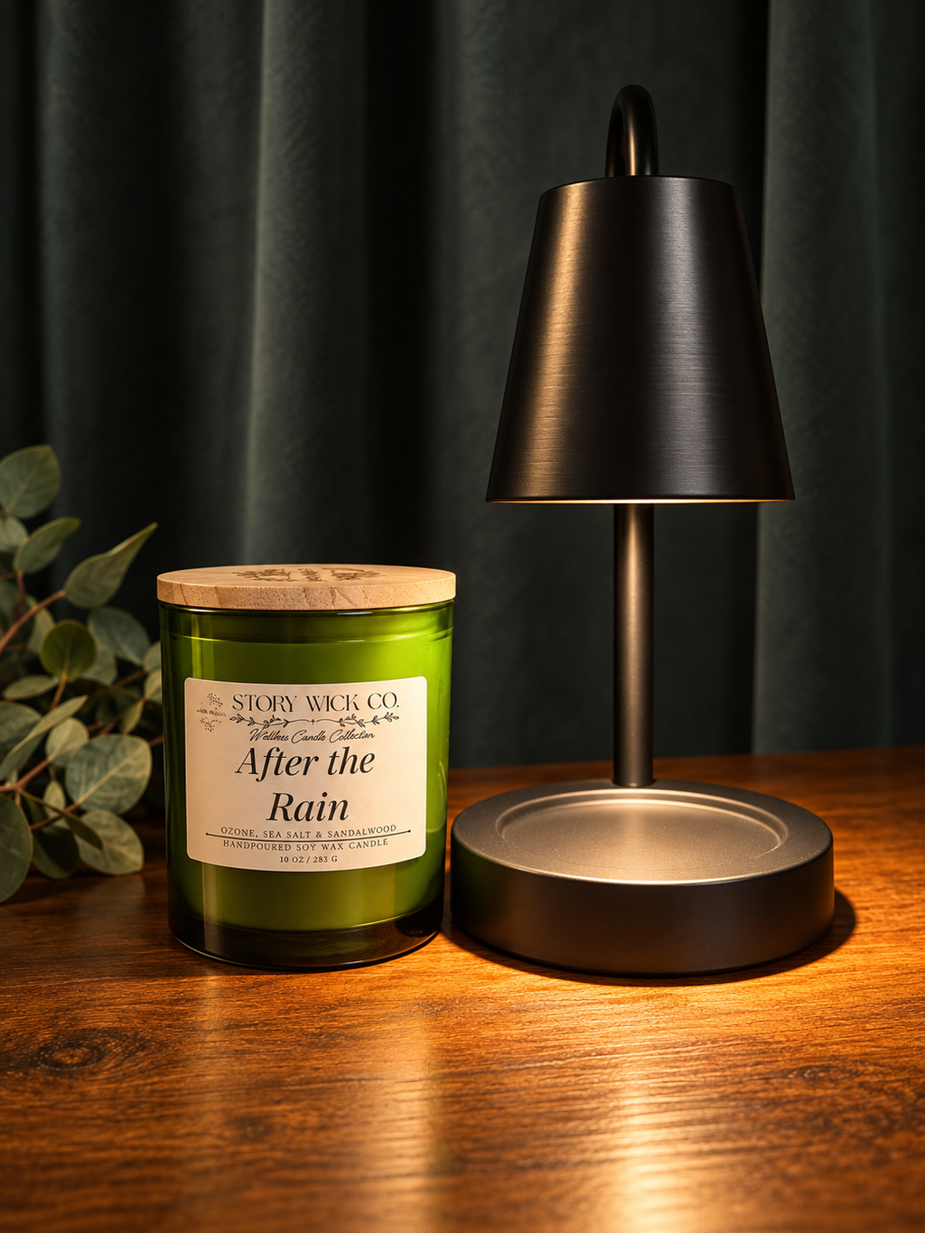A green glass jar candle labeled 'After the Rain' by Story Wick Co., styled on a wooden table next to a black table lamp against a dark curtain background.