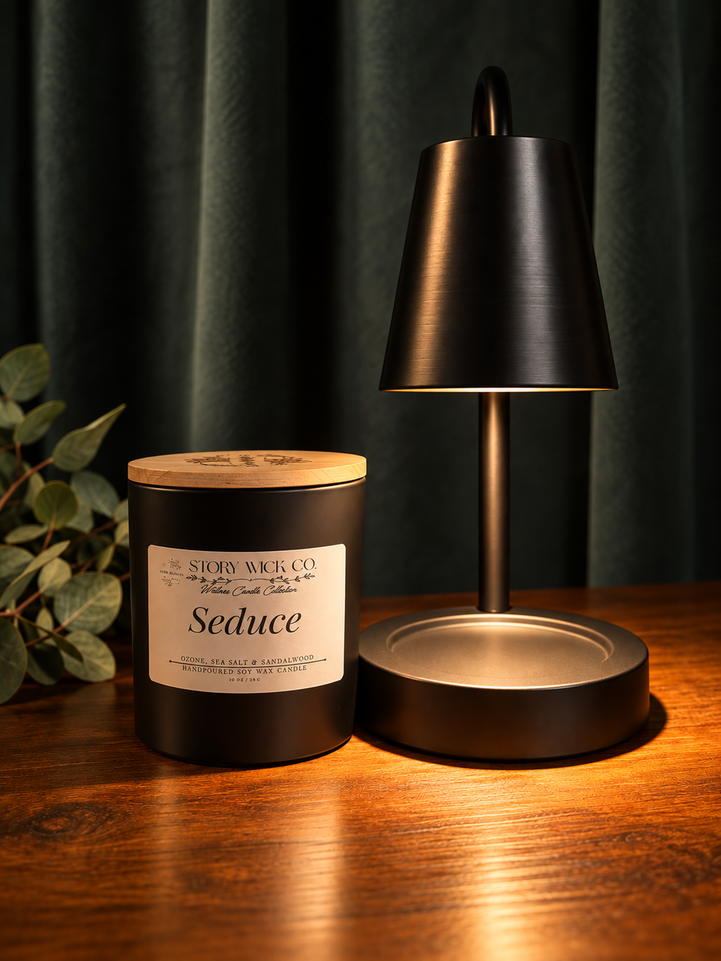 A dark metal scented candle jar labeled 'Seduce' by Story Wick Co sits on a wooden table next to a matching dark metal table lamp with a warm glowing shade, set against a dark green curtain background.