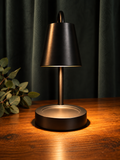A modern table lamp with a matte black metal shade and base sits on a warm wooden table, casting a soft glow upward. Dark green curtains hang in the background, with a small green plant visible on the left side.