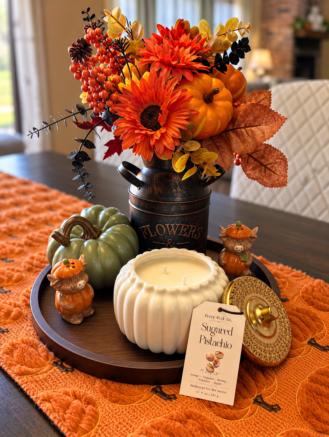 A styled autumn tabletop arrangement featuring a faux flower bouquet in a rustic metal canister, a white ribbed candle, a small green pumpkin, two orange pumpkin figurines, and a tray with a scent tag reading 'Sugared Pistachio' by Story Wick Co.