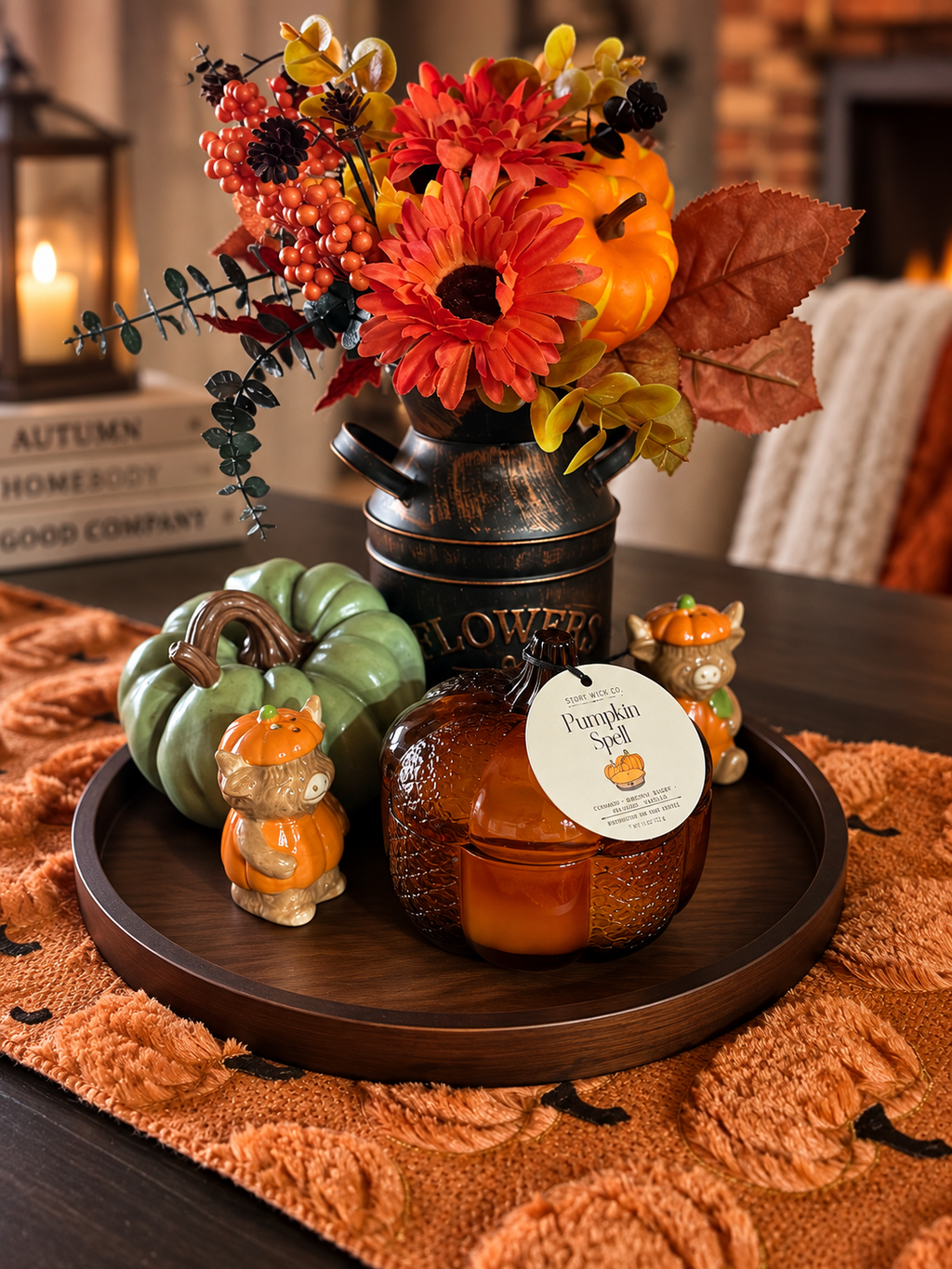An autumn-themed candle display on a dark wooden tray, featuring a pumpkin-shaped candle, a green ceramic pumpkin, and two small ceramic bears, styled with a vintage-style black metal flower pot filled with fall flowers and leaves.