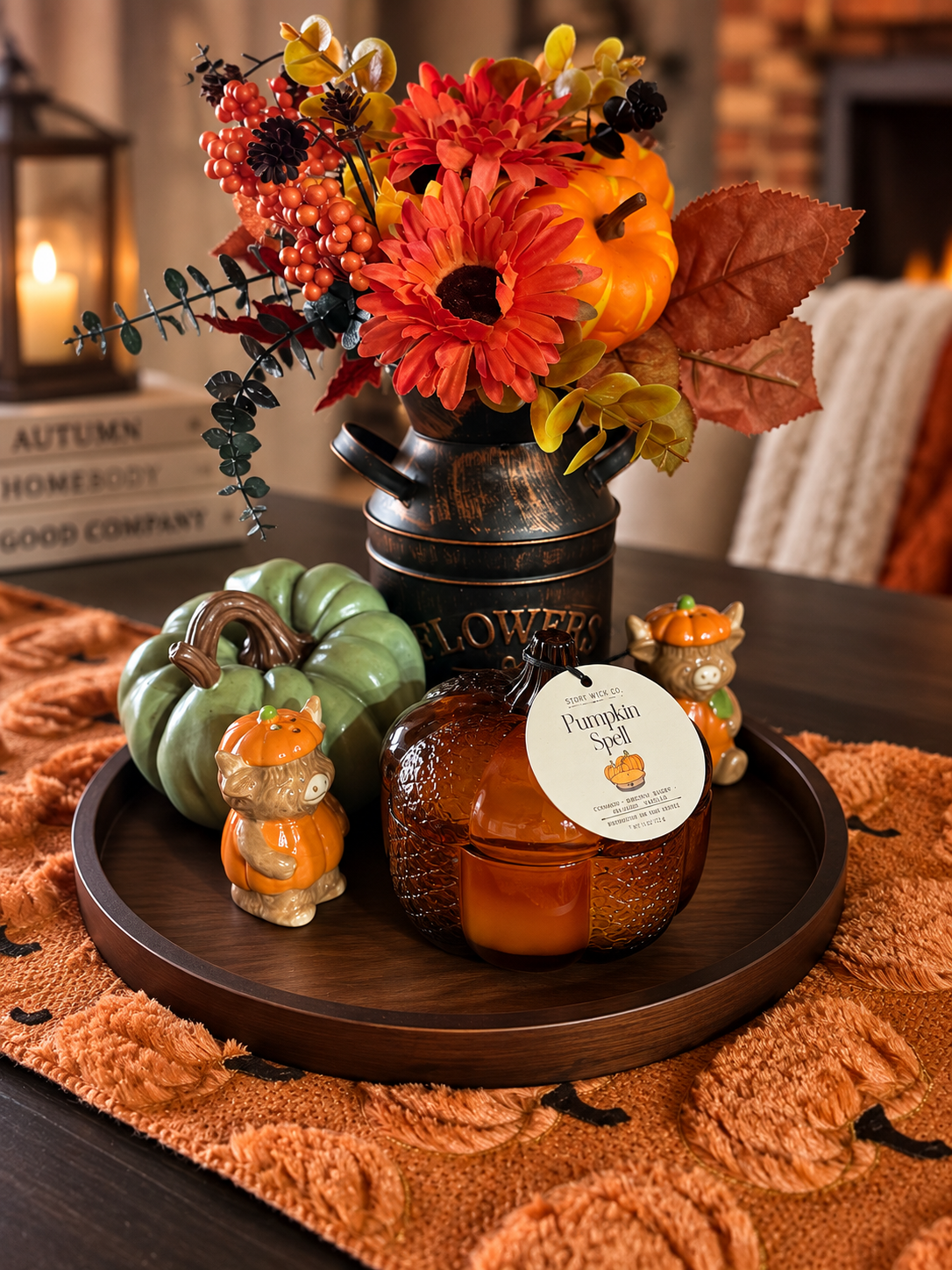 An autumn-themed candle display on a dark wooden tray, featuring a pumpkin-shaped candle, a green ceramic pumpkin, and two small ceramic bears, styled with a vintage-style black metal flower pot filled with fall flowers and leaves.