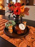 An autumn-themed tabletop centerpiece on a dark wood tray, featuring a faux flower arrangement in a dark metal pitcher, a candle in a glass jar, small decorative pumpkins, and a green pumpkin on an orange textured runner.