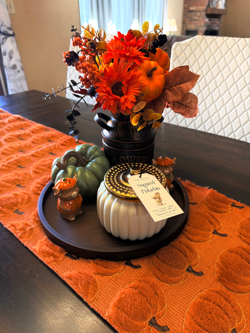 An autumn-themed tabletop centerpiece on a dark wood table, featuring a black metal bucket vase filled with orange and red artificial flowers, orange pumpkins, a small green pumpkin, a white ribbed pumpkin candle with a 'Sugared Pistachio' tag, and a small orange pumpkin figurine, all arranged on an orange textured runner.