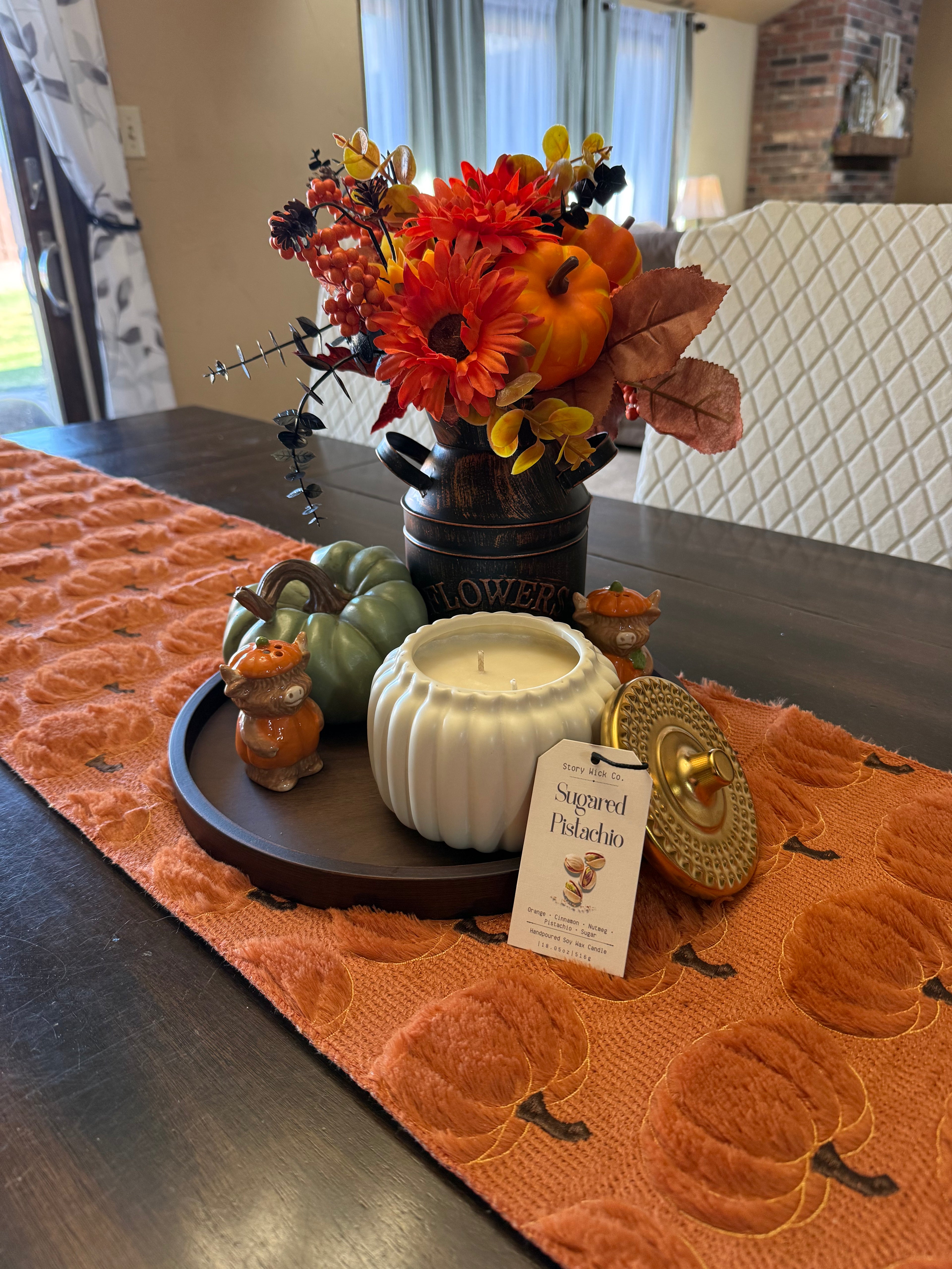 An autumn-themed table runner centerpiece set on a dark wood dining table, featuring a textured orange quilted runner, a black metal flower pot filled with fall flowers, a white ribbed candle, small decorative pumpkins, and a gold-toned lid.