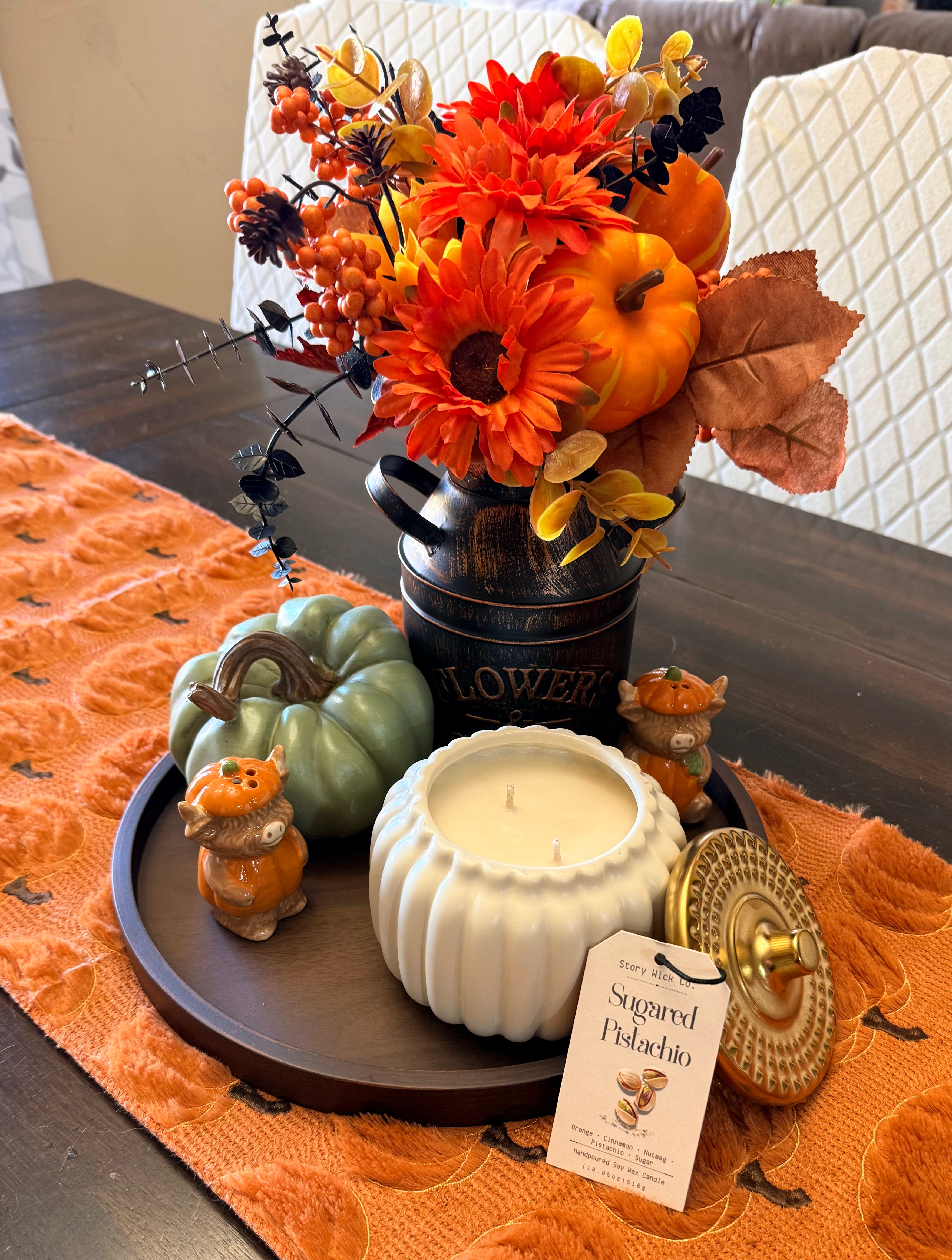 A styled autumn tabletop centerpiece featuring a faux floral arrangement in a dark metal flower pot, orange pumpkins, a sage green pumpkin, ceramic owl figurines, a white candle in a ribbed pumpkin-shaped holder, and a woven basket lid on an orange table runner.