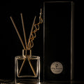 A clear glass reed diffuser bottle with metallic gold reeds stands beside a tall black box. The box has a round white label reading 'Misty Meadow' and 'Reed diffuser'.