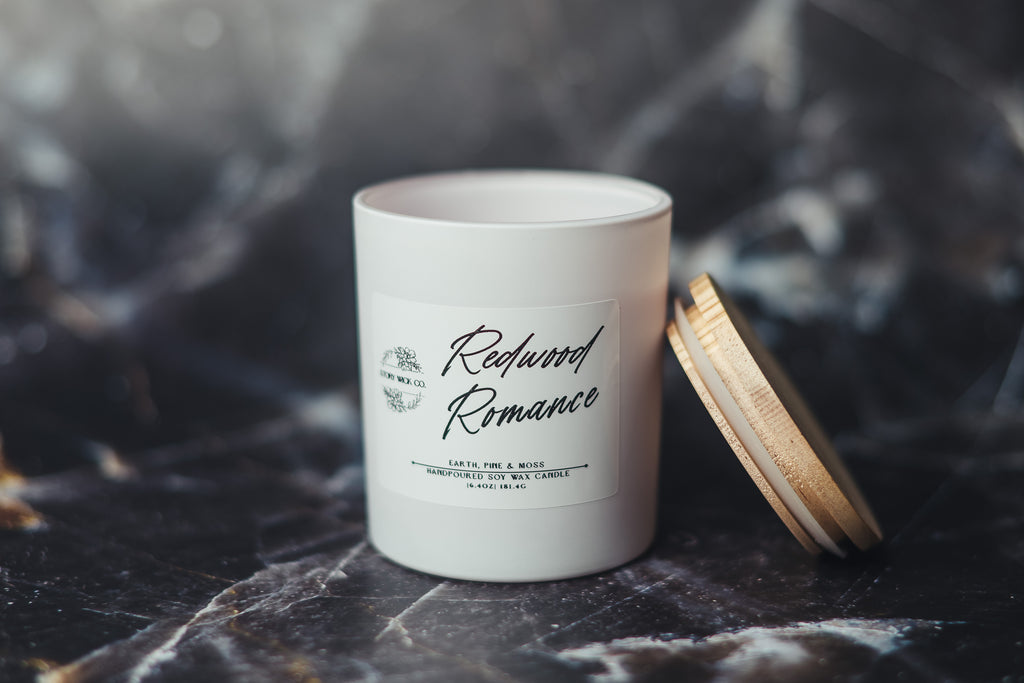 White candle labeled 'Redwood Romance' on a dark surface.