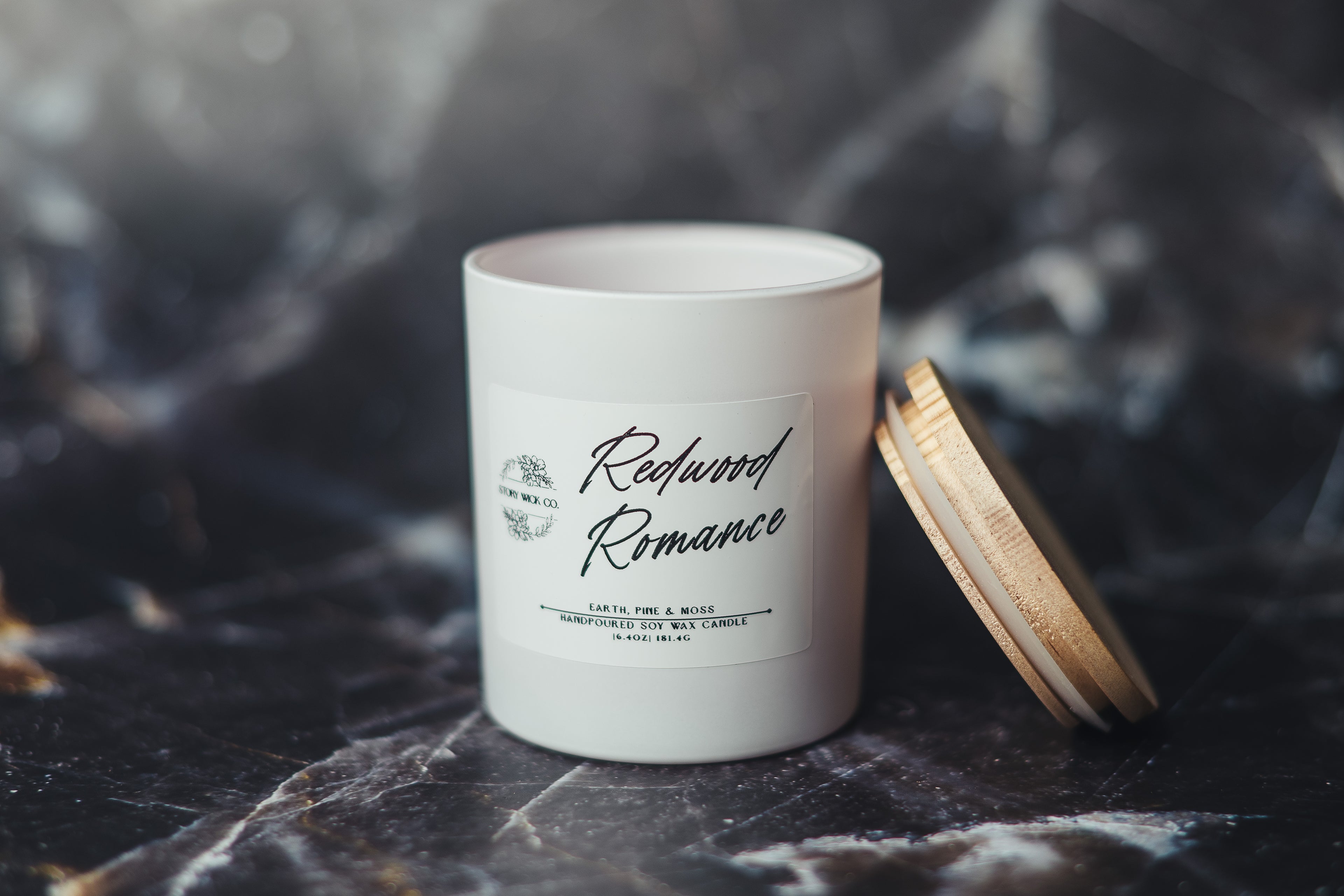 White candle labeled 'Redwood Romance' on a dark surface.