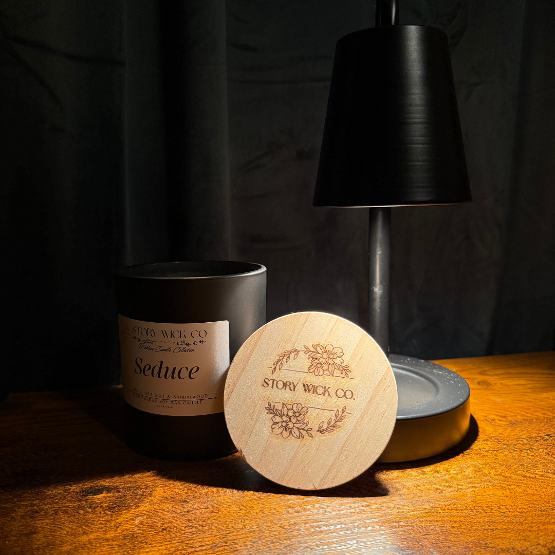 A candle jar with a matching round wooden lid placed in front of it on a wooden table. The candle container is matte black with a cream label reading 'Seduce' and 'Story Wick Co.
