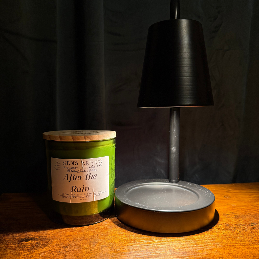 A green candle jar with a wooden lid and white label reading Story Wick Co, 'After the Rain' sits on a wooden table beside a black table lamp in a dimly lit setting.