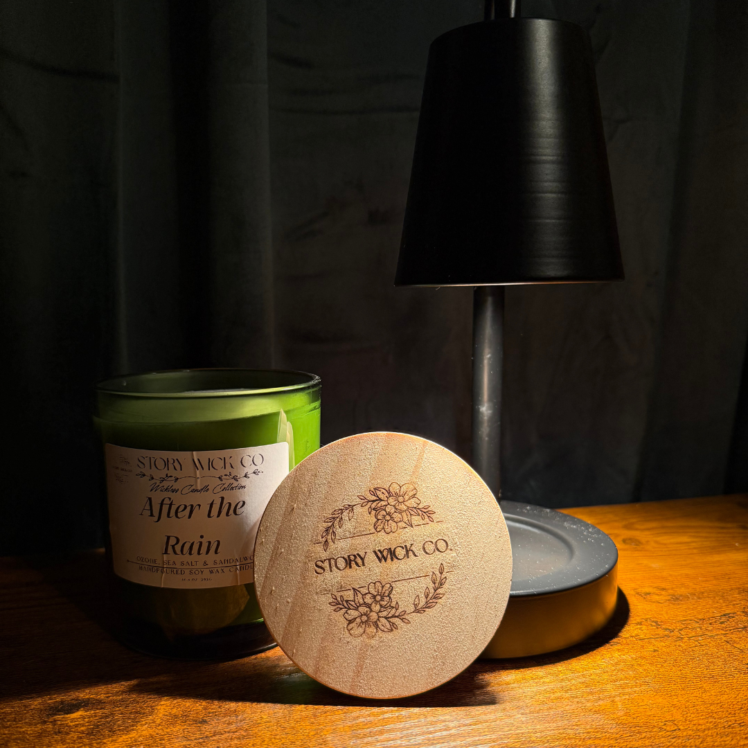 A wooden round coaster or lid engraved with 'STORY WICK CO.' and floral motifs, placed in front of a green candle jar labeled 'After the Rain' on a wooden table, with a black lamp in the background.