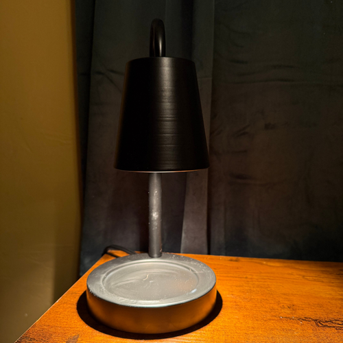 A modern table lamp with a matte black tapered shade and a brushed silver metal base, shown lit on a wooden surface against a dark backdrop.