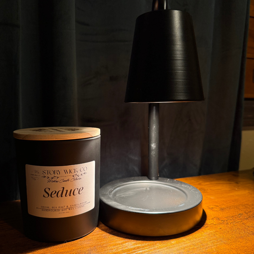 A dark brown candle jar with a light wood lid and a white label reading 'Seduce' by Story Wick Co, placed on a wooden table next to a black table lamp against a dark curtain background.