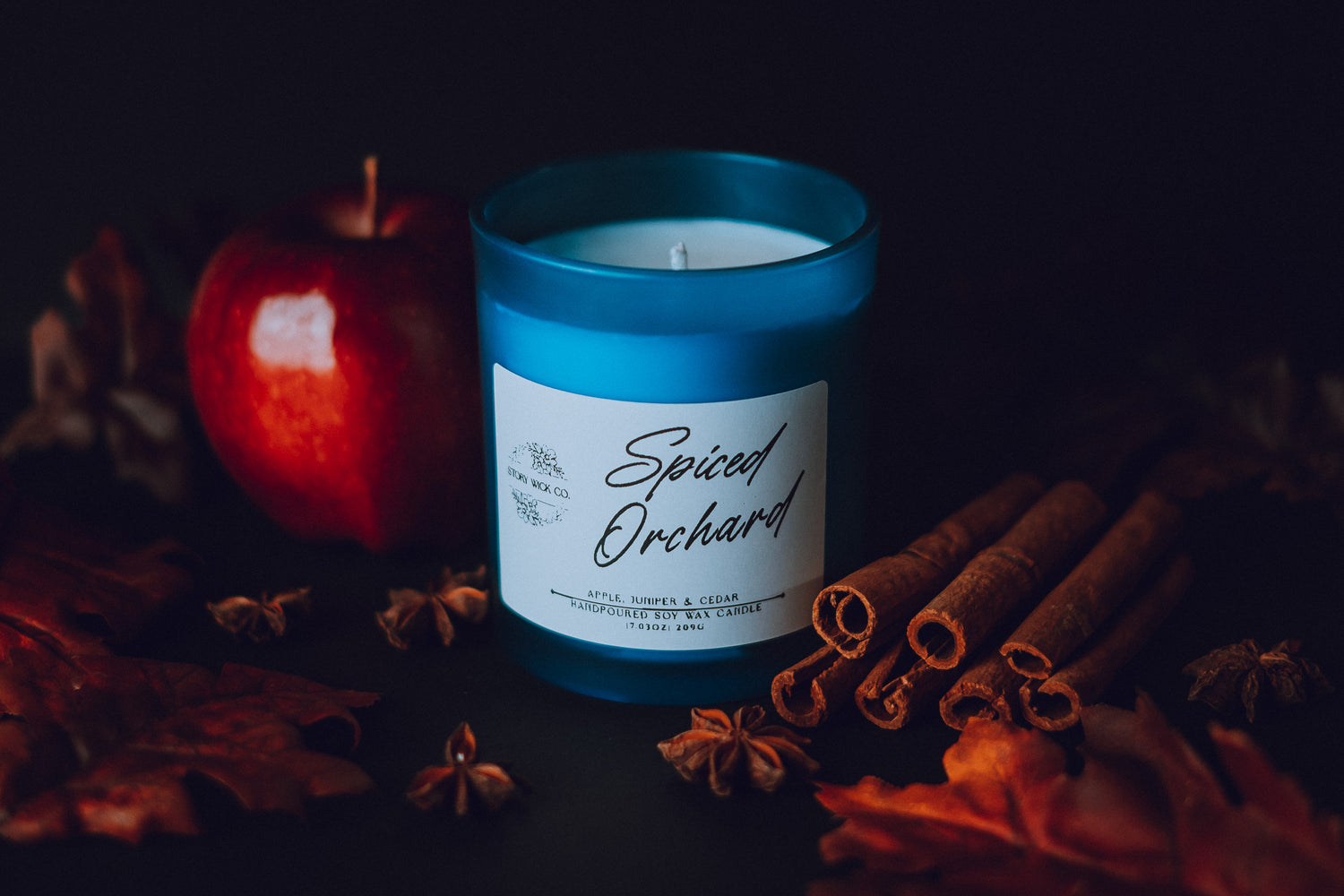 Candle labeled 'Spiced Orchard' with an apple, cinnamon sticks, and star anise on a dark background