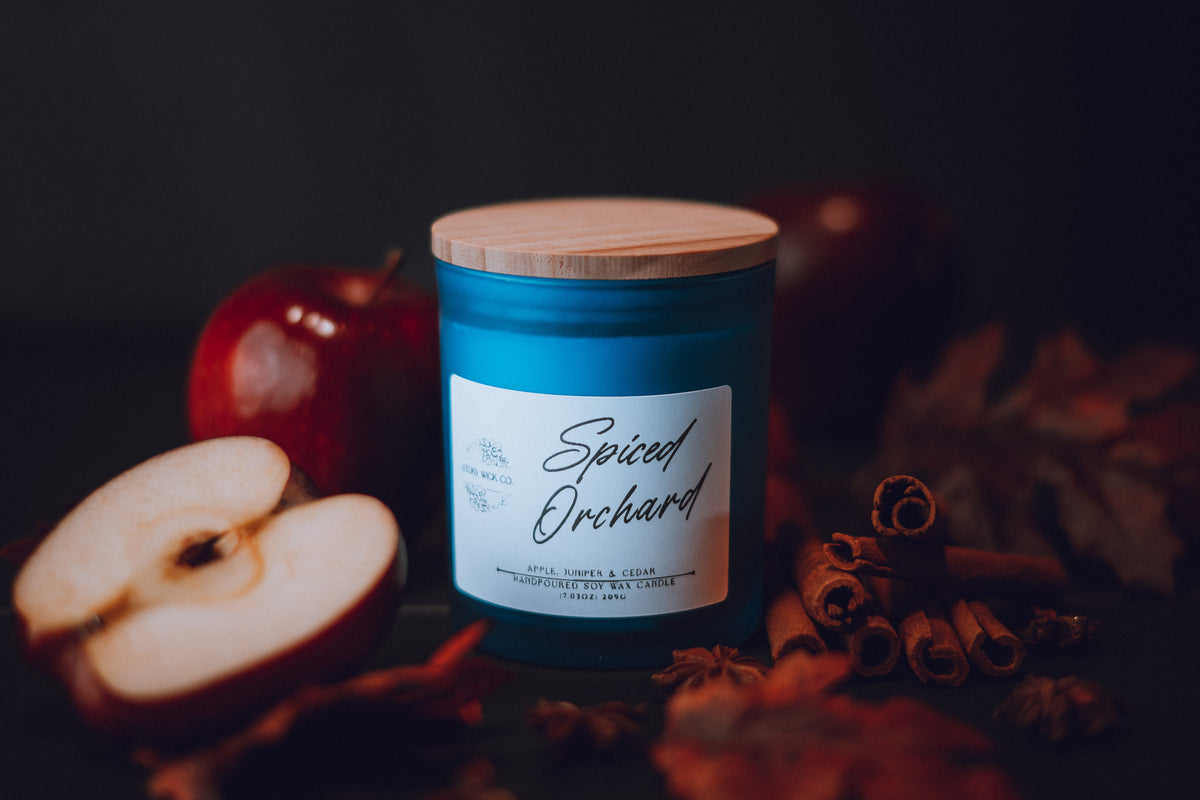 Candle labeled 'Spiced Orchard' with apples and cinnamon sticks on a dark background.