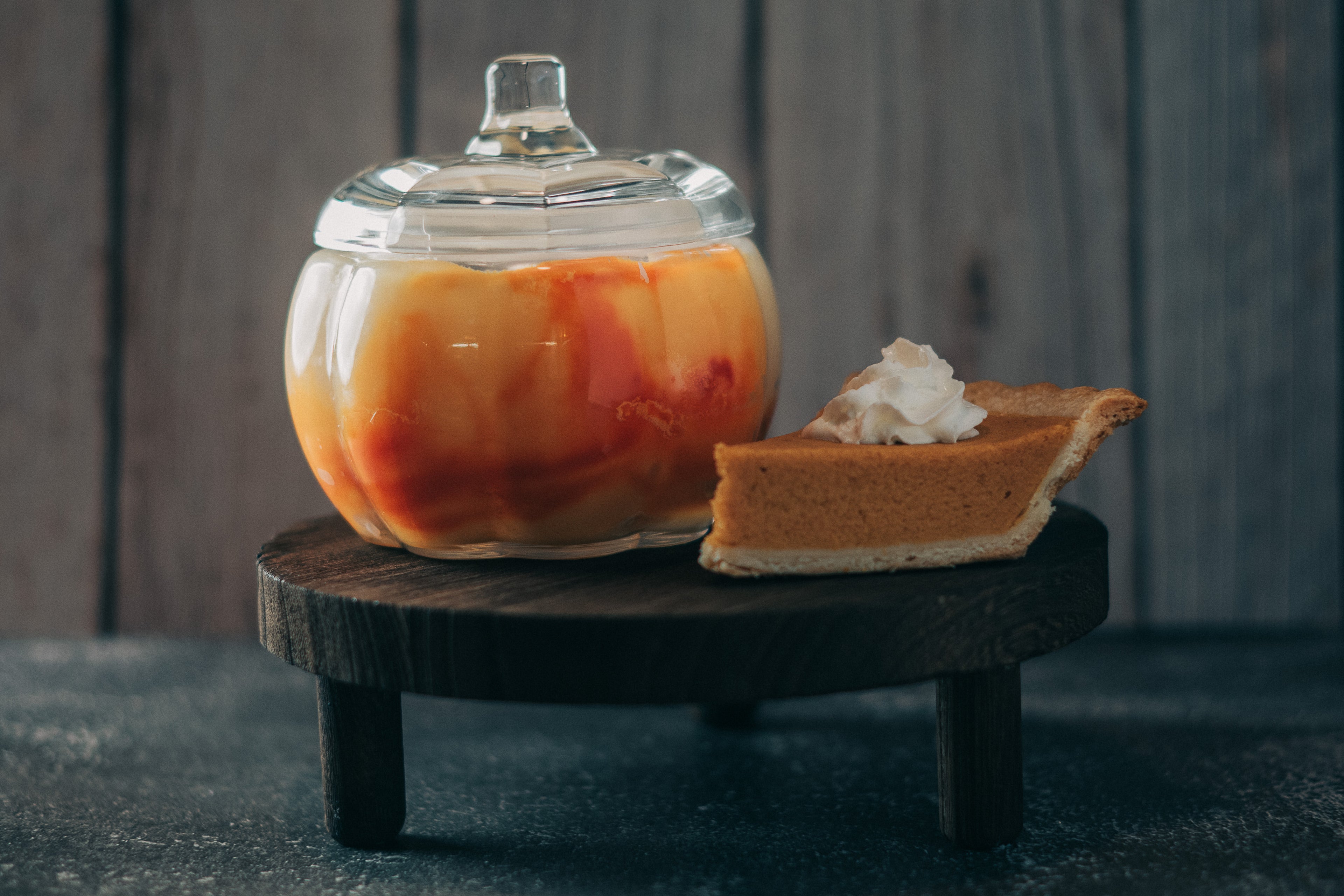 Glass container with a red and orange pumpkin shaped candle on a wooden stand with a slice of pie.